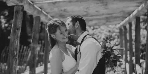 Black and white photograph of a bride and groom embracing under a wooden pergola structure.