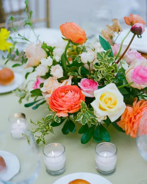 A colorful floral centerpiece featuring orange ranunculus, white and pink roses, and greenery arranged on a dining table with lit candles.