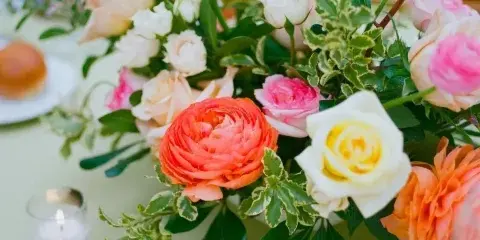 A colorful floral centerpiece featuring orange ranunculus, white and pink roses, and greenery arranged on a dining table with lit candles.