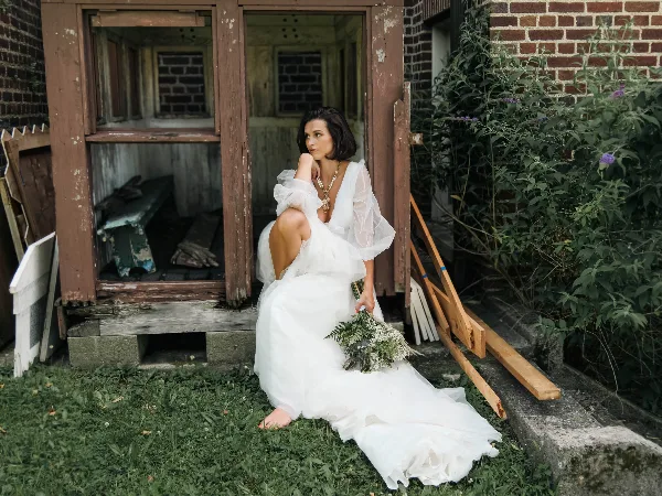 A bride in a white gown sitting on the grass in front of a rustic wooden structure, holding a bouquet.