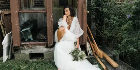A bride in a white gown sitting on the grass in front of a rustic wooden structure, holding a bouquet.