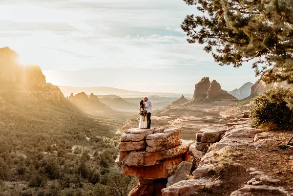 A bride and groom standing on a rocky outcrop overlooking a vast desert landscape with red rock formations in the background.
