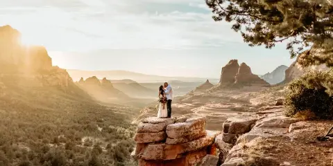 A bride and groom standing on a rocky outcrop overlooking a vast desert landscape with red rock formations in the background.