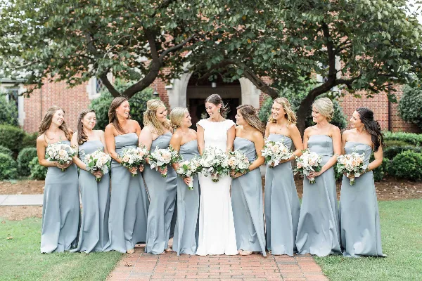 A bride and bridesmaids in grey dresses holding matching white and peach floral bouquets outdoors.