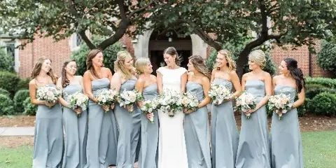 A bride and bridesmaids in grey dresses holding matching white and peach floral bouquets outdoors.