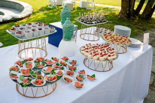 An outdoor catering buffet table featuring various appetizers, canapés, and food displays arranged on tiered stands.