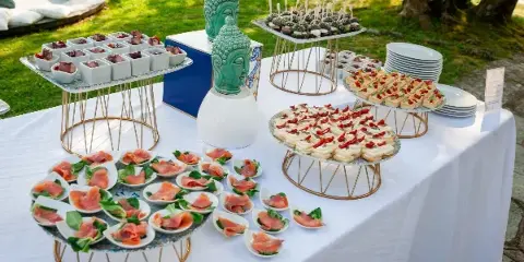 An outdoor catering buffet table featuring various appetizers, canapés, and food displays arranged on tiered stands.