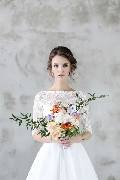 A bride in a white lace dress holding a colorful floral bouquet with greenery.