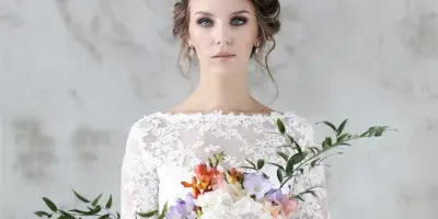 A bride in a white lace dress holding a colorful floral bouquet with greenery.