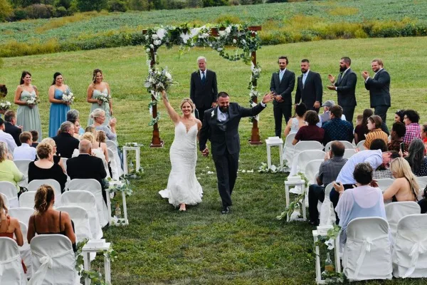 A bride and groom walking down the aisle during an outdoor wedding ceremony with guests seated in white chairs.