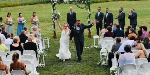 A bride and groom walking down the aisle during an outdoor wedding ceremony with guests seated in white chairs.