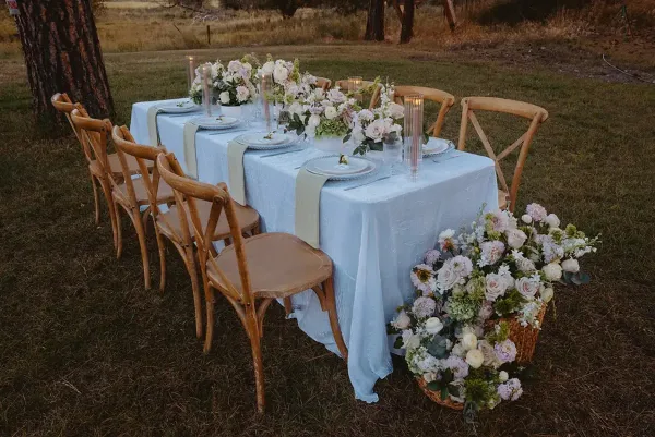 A long outdoor reception table set with white linens, crossback chairs, and floral centerpieces under pine trees.