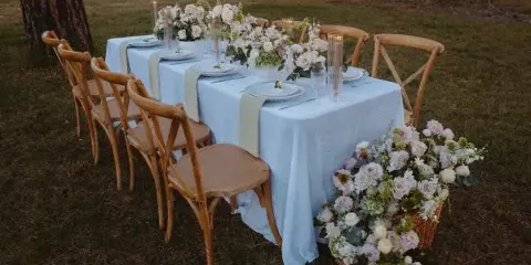 A long outdoor reception table set with white linens, crossback chairs, and floral centerpieces under pine trees.