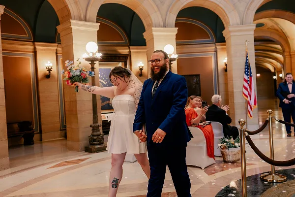 A bride and groom walking hand-in-hand through a grand, arched interior hall during their wedding.