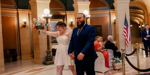 A bride and groom walking hand-in-hand through a grand, arched interior hall during their wedding.