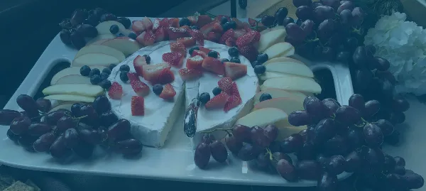 A catering platter featuring a large wheel of soft cheese topped with strawberries and blueberries, surrounded by apple slices and red grapes.