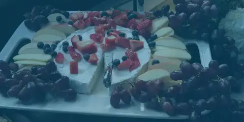 A catering platter featuring a large wheel of soft cheese topped with strawberries and blueberries, surrounded by apple slices and red grapes.