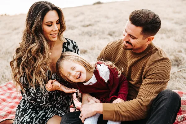 A family of three sitting on a red checkered blanket in a dry grassy field, with the mother and father smiling at their laughing daughter.