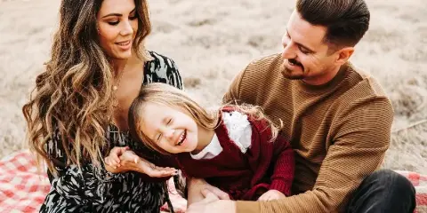 A family of three sitting on a red checkered blanket in a dry grassy field, with the mother and father smiling at their laughing daughter.
