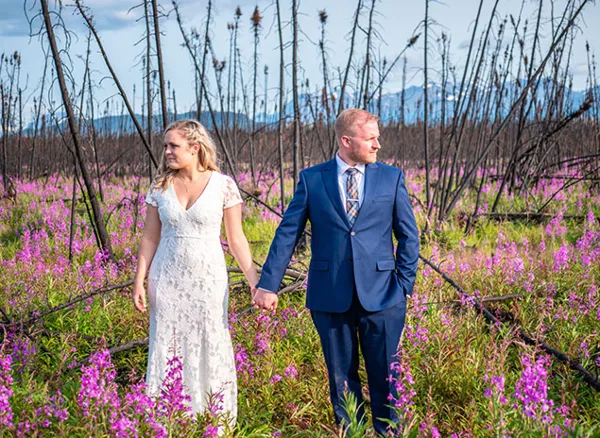 A bride and groom holding hands while walking through a field of purple wildflowers.