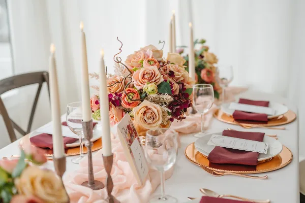 A close-up of an elegant wedding table setting featuring a floral centerpiece with peach roses, tall white candles in brass holders, gold charger plates, and burgundy napkins.