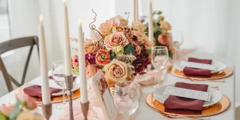 A close-up of an elegant wedding table setting featuring a floral centerpiece with peach roses, tall white candles in brass holders, gold charger plates, and burgundy napkins.