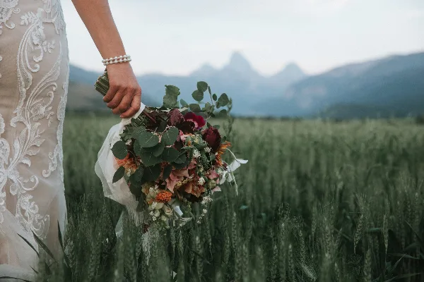 A close-up of a bride's hand holding a floral bouquet in a field with mountains in the background.