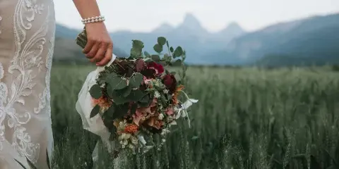 A close-up of a bride's hand holding a floral bouquet in a field with mountains in the background.