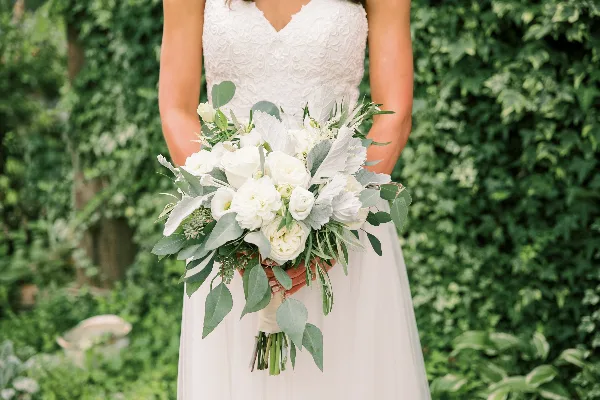 A bride in a white lace dress holding a white floral bouquet with greenery against a leafy background.