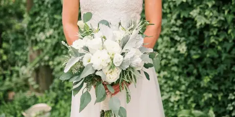 A bride in a white lace dress holding a white floral bouquet with greenery against a leafy background.