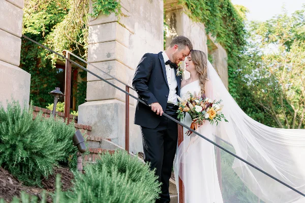 A bride and groom standing close together on stone steps outdoors, with the bride holding a bouquet and wearing a flowing veil.