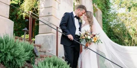 A bride and groom standing close together on stone steps outdoors, with the bride holding a bouquet and wearing a flowing veil.
