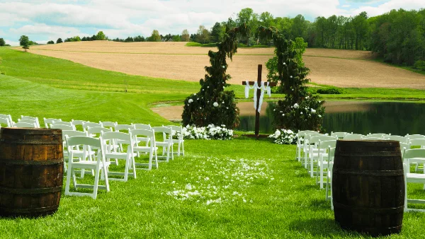 An outdoor wedding ceremony setup with white chairs, a floral arch, and a grassy aisle leading to a pond.