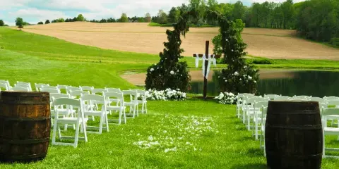 An outdoor wedding ceremony setup with white chairs, a floral arch, and a grassy aisle leading to a pond.