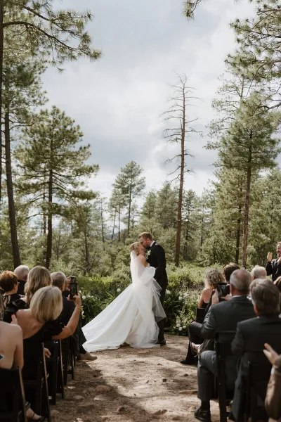 A bride and groom sharing a kiss during an outdoor wedding ceremony surrounded by tall pine trees and seated guests.