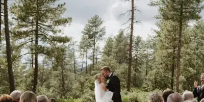 A bride and groom sharing a kiss during an outdoor wedding ceremony surrounded by tall pine trees and seated guests.