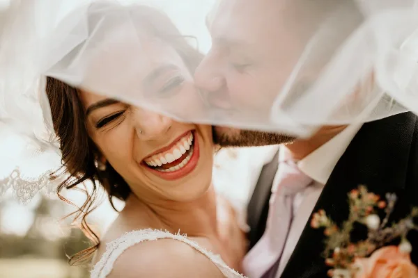Bride with styled hair and makeup smiling under a veil next to a groom in a wedding suit.