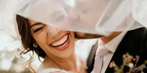 Bride with styled hair and makeup smiling under a veil next to a groom in a wedding suit.