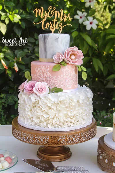 A three-tier custom wedding cake with white ruffles, pink lace, and fresh flowers on a gold stand.