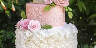 A three-tier custom wedding cake with white ruffles, pink lace, and fresh flowers on a gold stand.