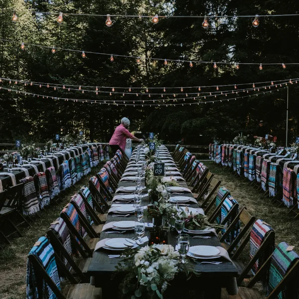 An outdoor wedding reception setup featuring a long wooden farm table, rows of chairs with blankets, and overhead string lights.