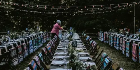 An outdoor wedding reception setup featuring a long wooden farm table, rows of chairs with blankets, and overhead string lights.