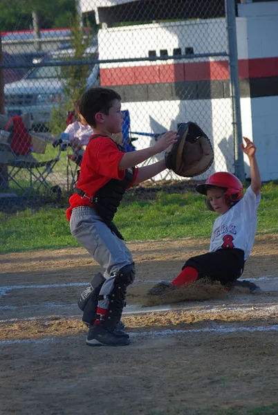 A youth baseball player in a red jersey and catcher's gear catches a ball while another player slides into base.