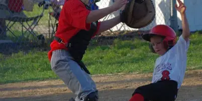 A youth baseball player in a red jersey and catcher's gear catches a ball while another player slides into base.