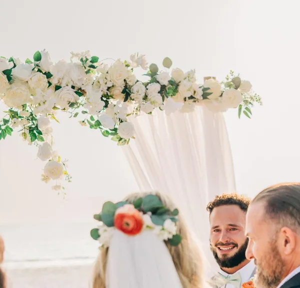 A bride wearing a flower crown and a groom standing under a white floral arch during a beach wedding ceremony.