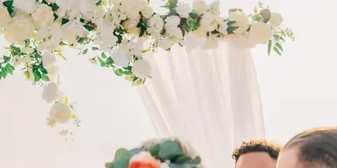 A bride wearing a flower crown and a groom standing under a white floral arch during a beach wedding ceremony.
