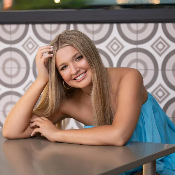 A smiling young woman with long blonde hair and professional makeup, wearing a blue dress, posing in front of a patterned tile wall.
