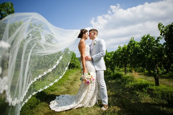 A bride and groom standing close together in a vineyard, with the bride's veil blowing in the wind.