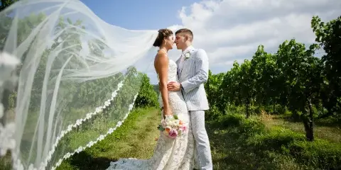 A bride and groom standing close together in a vineyard, with the bride's veil blowing in the wind.