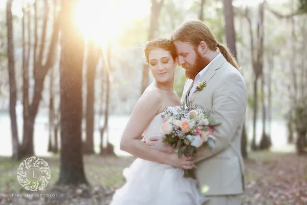 A bride and groom embracing in a wooded area near a river, with the bride holding a bouquet.
